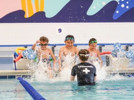 Three young swimmers sit on the edge of a pool laughing and kicking their feet, splashing their instructor