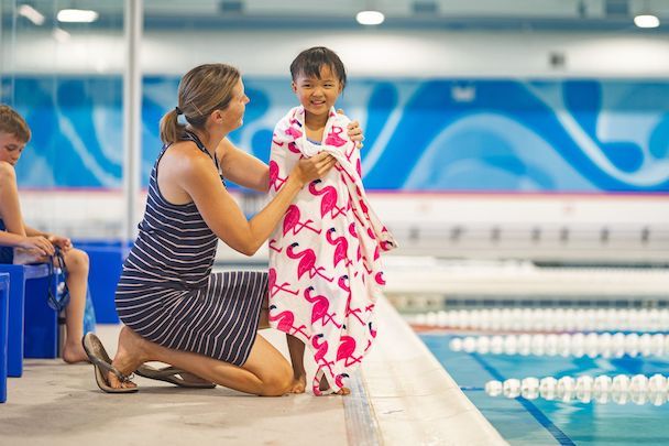 A young swimmer wrapped in a towel smiles as her guardian helps to dry her off on the pool deck