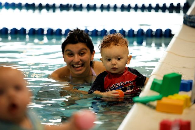 A little baby is being accompanied by his mom, who is supporting him to overcome his fear of water