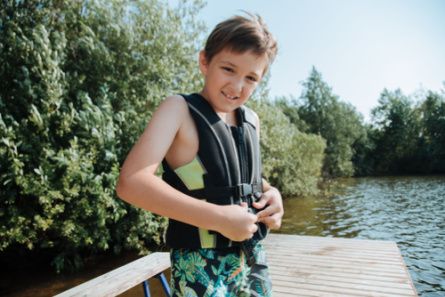A young boy in a life vest on a dock at a lake smiles at the camera