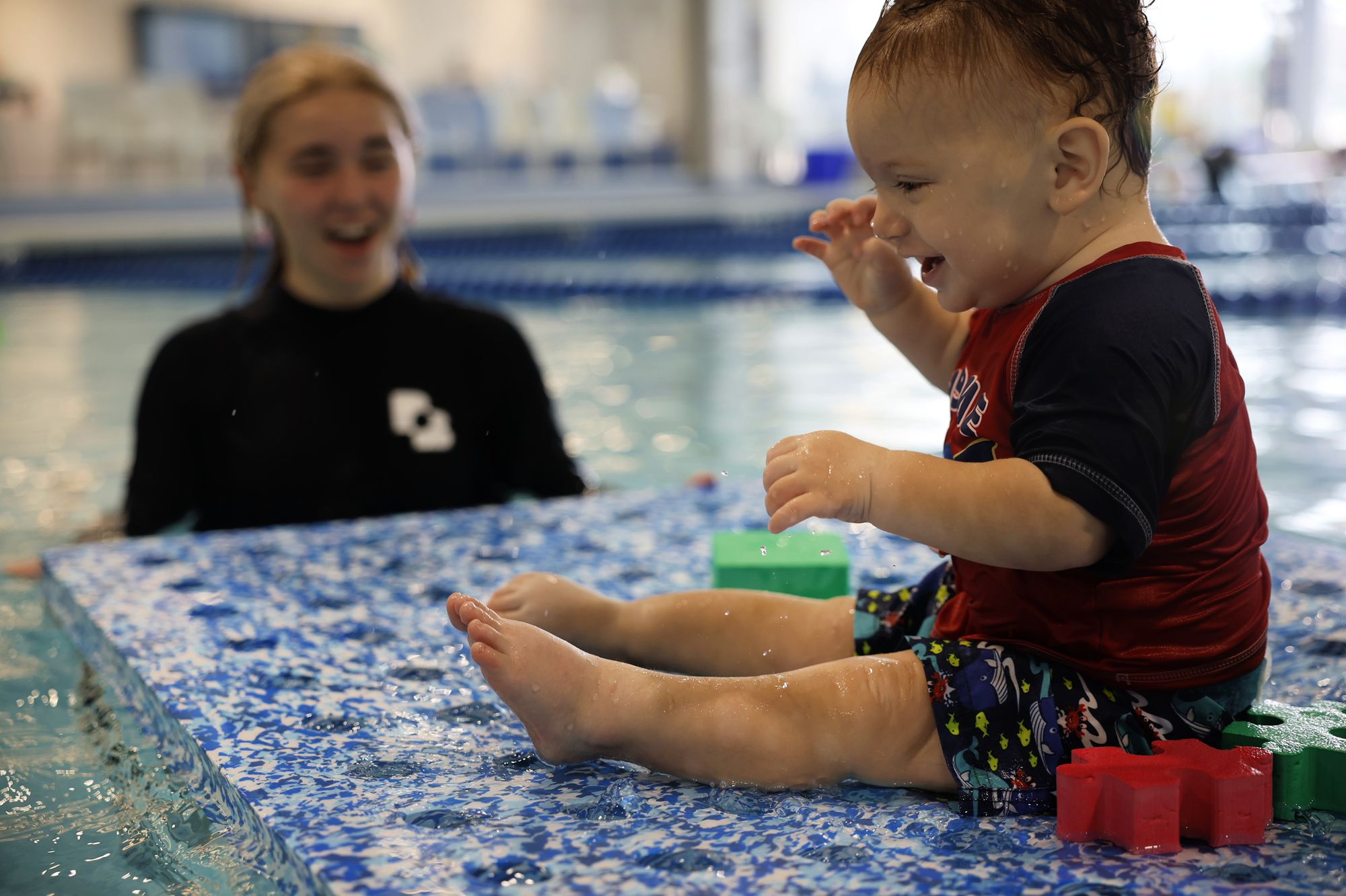 Small baby sits on a floating mat in a pool with swim toys with swim instructor