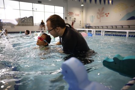 A young swimmer with pink goggles is guided through the pool by her instructor 