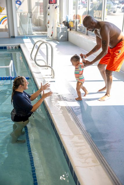 A baby is walking, being led by her father, towards the pool and swim instructor at Big Blue Swim School