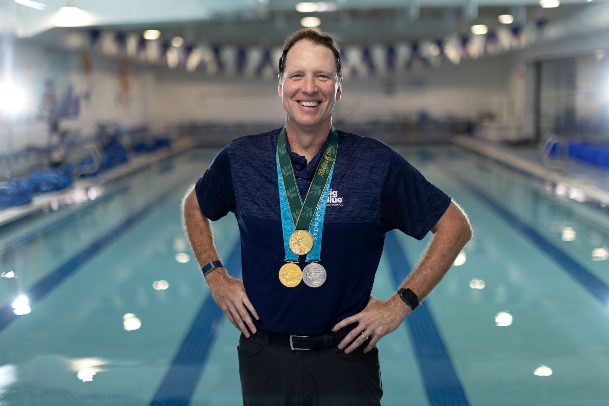 Olympic medalist, Tom Dolan, stands on a Big Blue pool deck, wearing his medals