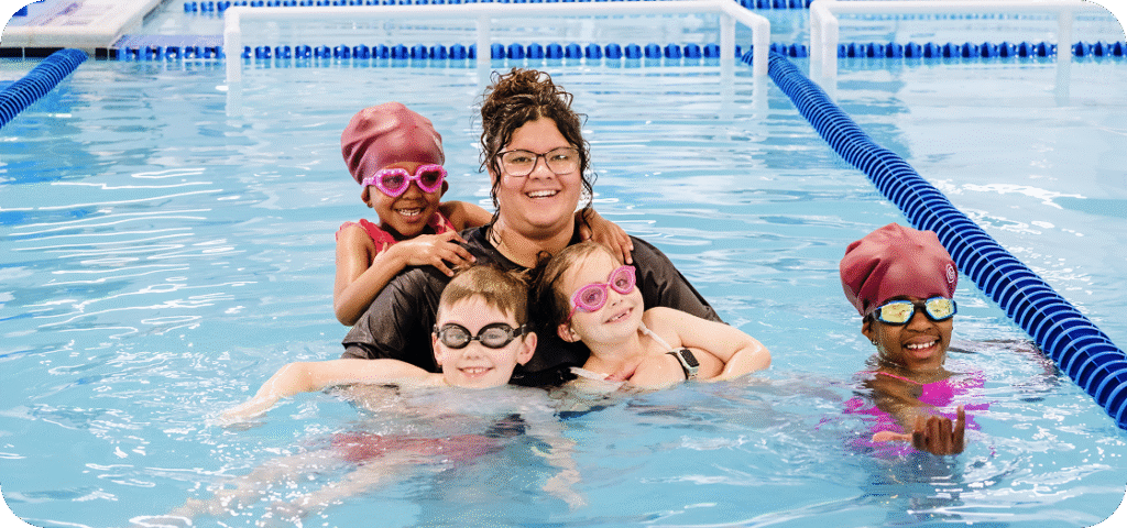 A swimming instructor and four children in a pool, all wearing goggles and swim caps, smiling at the camera.