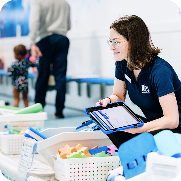 A woman in a navy "Big Blue" shirt holds a tablet and kneels near bins of pool toys; a child and adult are visible in the background.