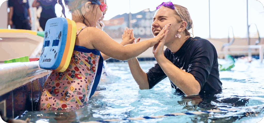 A young girl in a patterned swimsuit and a swim float, wearing pink goggles, high-fives a smiling swim instructor wearing purple goggles and a black swim shirt in a swimming pool.