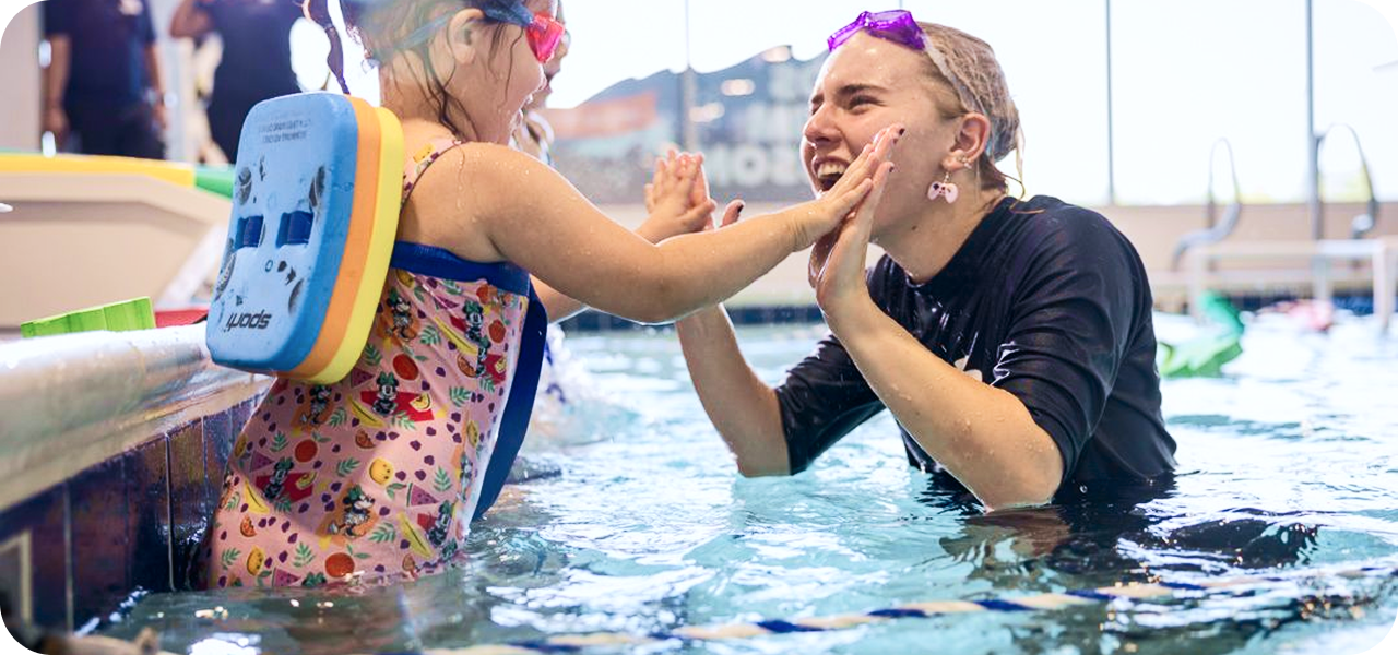 A young girl in a patterned swimsuit and a swim float, wearing pink goggles, high-fives a smiling swim instructor wearing purple goggles and a black swim shirt in a swimming pool.