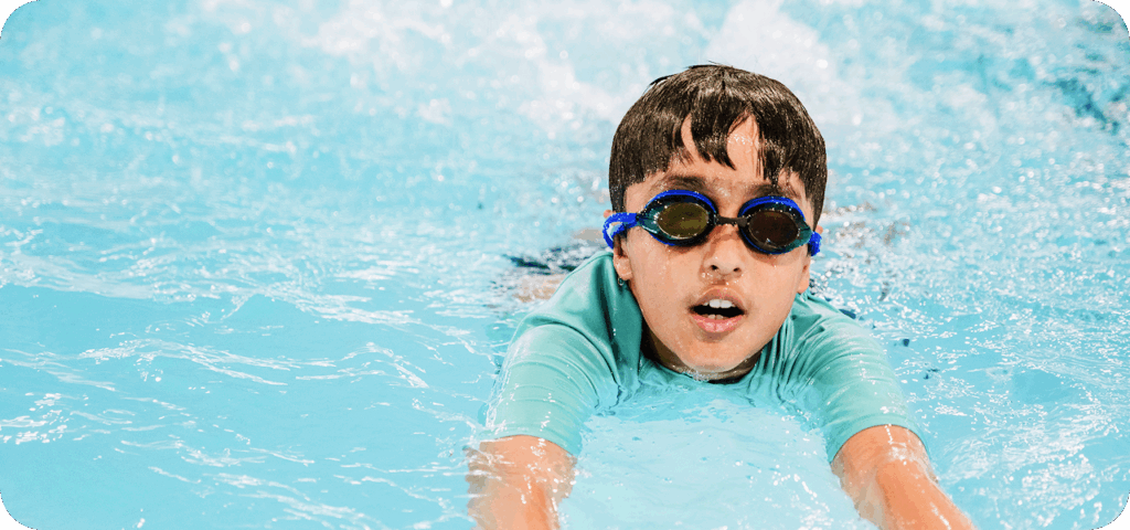 A young boy with dark hair and blue-tinted swim goggles swims freestyle in a pool, facing the viewer with an open mouth and slightly strained expression.