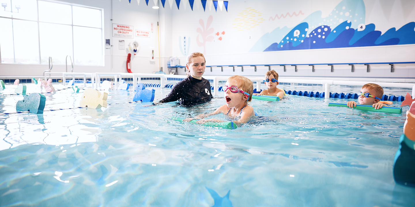 A swimming instructor in a black top assists three young children in a swimming lesson; the children wear goggles and use floatation devices.