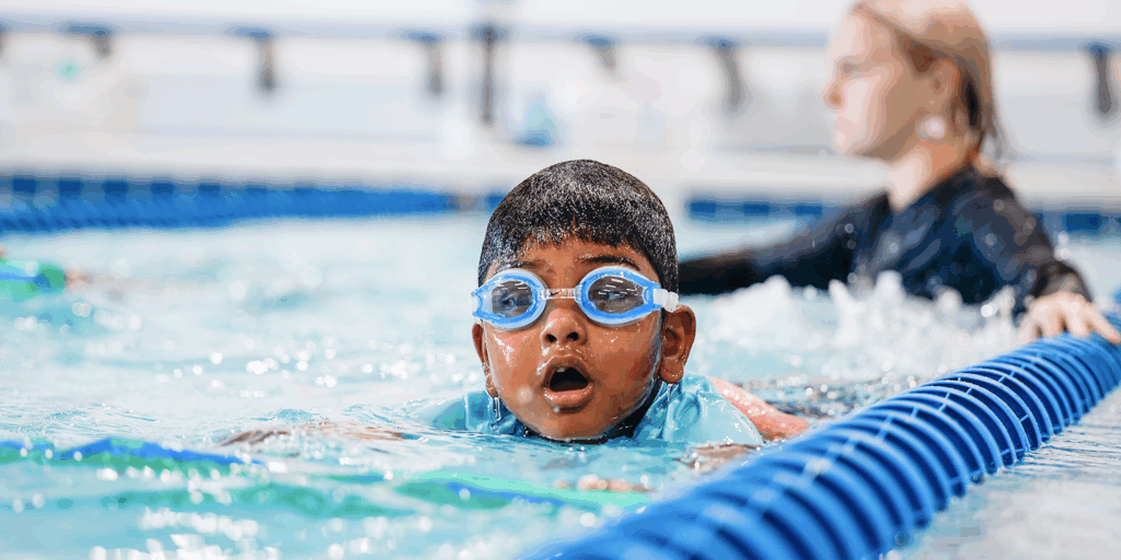 Young boy with goggles swimming freestyle in a pool, mouth open, with a swimming instructor visible in the background.