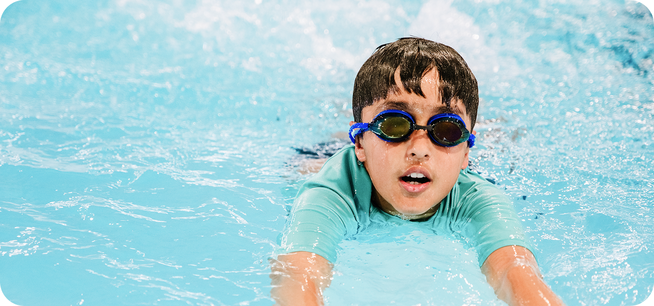 A young boy with dark hair and blue-tinted swim goggles swims freestyle in a pool, facing the viewer with an open mouth and slightly strained expression.
