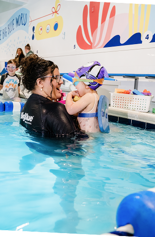 A woman in a black swimsuit, wearing glasses, and smiling, holds a young child in a swimming pool. The child wears a swim cap and goggles, a flotation device on their back, and holds a small yellow item.