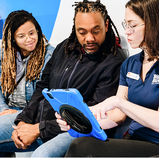 A woman in a blue shirt with a name tag labeled "Bank" points to a blue tablet screen held by a man with dreadlocks, while another woman with dreadlocks looks on.
