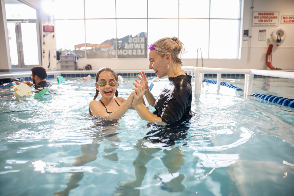 A young girl with pigtails and swim goggles smiles widely in a swimming pool, holding hands with a smiling swimming instructor who is wearing a black shirt and pink earrings.