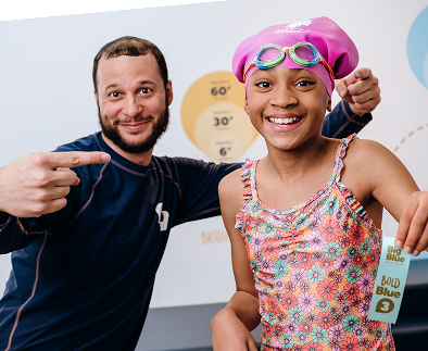 Smiling girl in a floral swimsuit and pink swim cap with goggles, holding a "Bold Blue 3" achievement ribbon, poses next to a smiling instructor wearing a dark blue shirt, both in a swimming lesson setting.