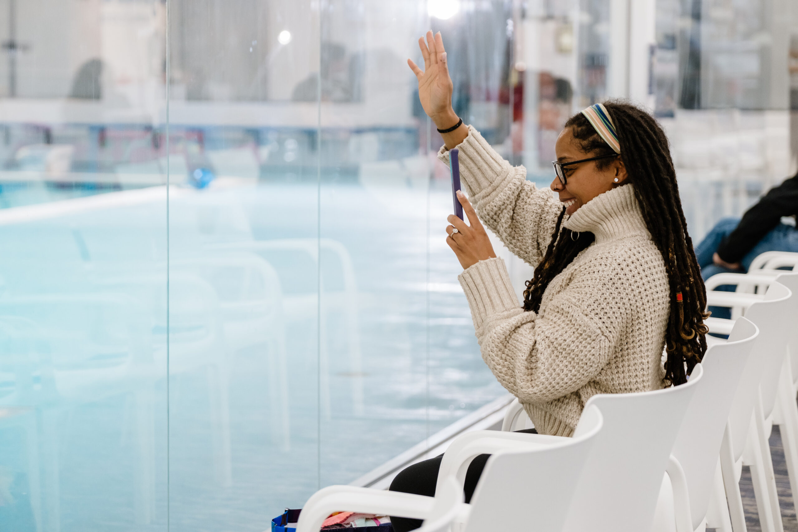 Woman with long dreadlocks wearing glasses and a headband, sitting in a white chair, holding a phone with one hand and waving with the other, smiling.