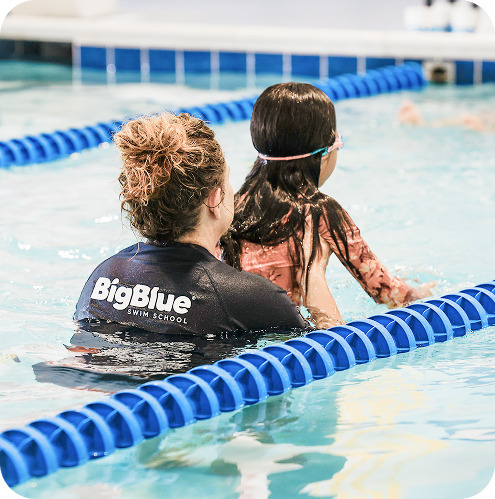 Female swim instructor in black shirt with Big Blue Swim School logo holding a young girl in a floral rash guard in a swimming pool, teaching her to swim.
