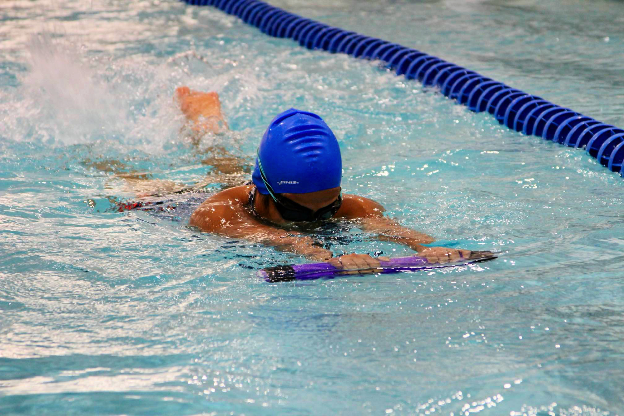 Swimmer in blue cap and goggles, using a purple kickboard, swimming in a pool.