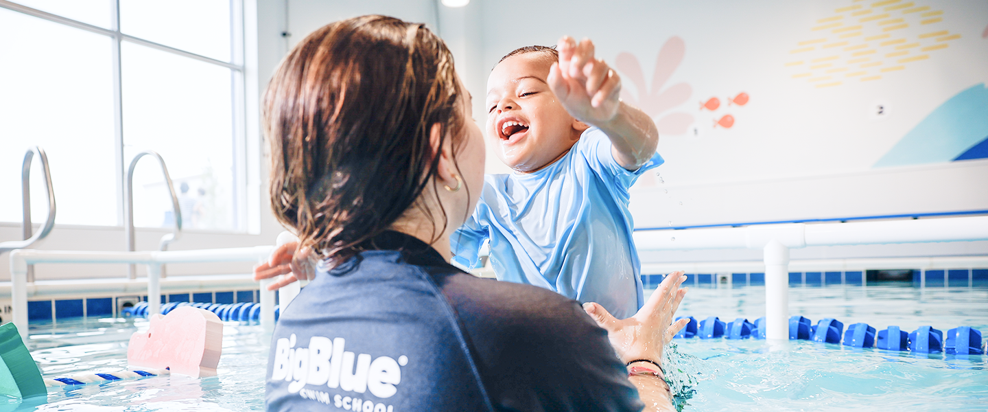A happy toddler wearing a blue swim shirt reaching towards a swimming instructor in a pool, at the Big Blue Swim School, with a smiling face and arms outstretched.
