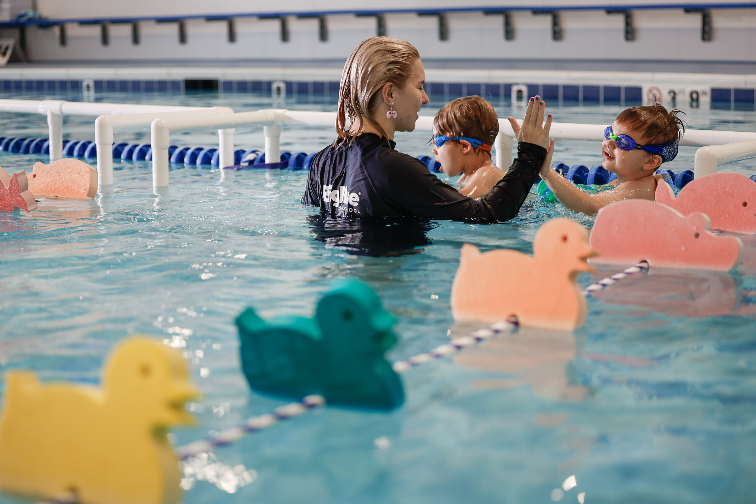 A blonde swim instructor in a black rash guard high-fives two young boys in goggles in a swimming pool, with duck-shaped floatation devices in the foreground.