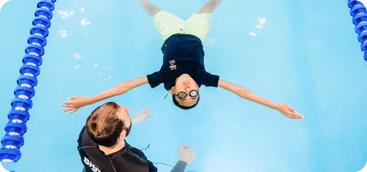 A young boy wearing swim goggles and a navy t-shirt floats face up in a swimming pool, arms outstretched to the sides. A swim instructor in a black shirt appears to be supporting him from below, and the blue water reflects the sky, with lane markers visible on the sides.