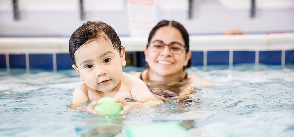 A young child, with dark wet hair and a curious expression, holds a green pool float while submerged in water. A woman wearing glasses smiles in the background, also in the water.