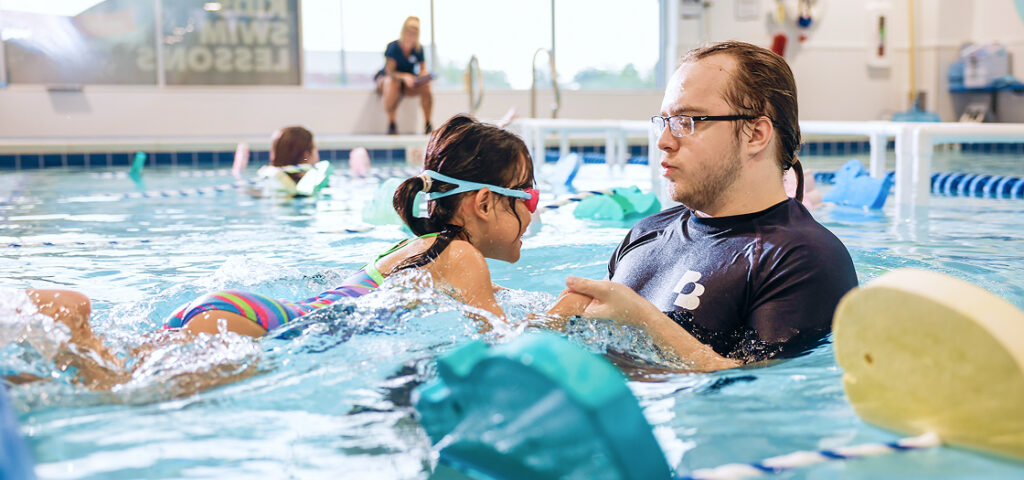 Young girl learning to swim in a pool with an instructor. The girl is in the water, wearing goggles and a swimsuit, and is mid-stroke, with water splashing around her. An instructor in a black shirt is next to her in the water, helping and guiding her.