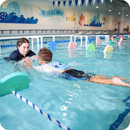 A young boy swims freestyle in a pool with an instructor, wearing a flotation device and goggles, while the instructor guides his arms; decorative foam animal shapes float in the lanes.