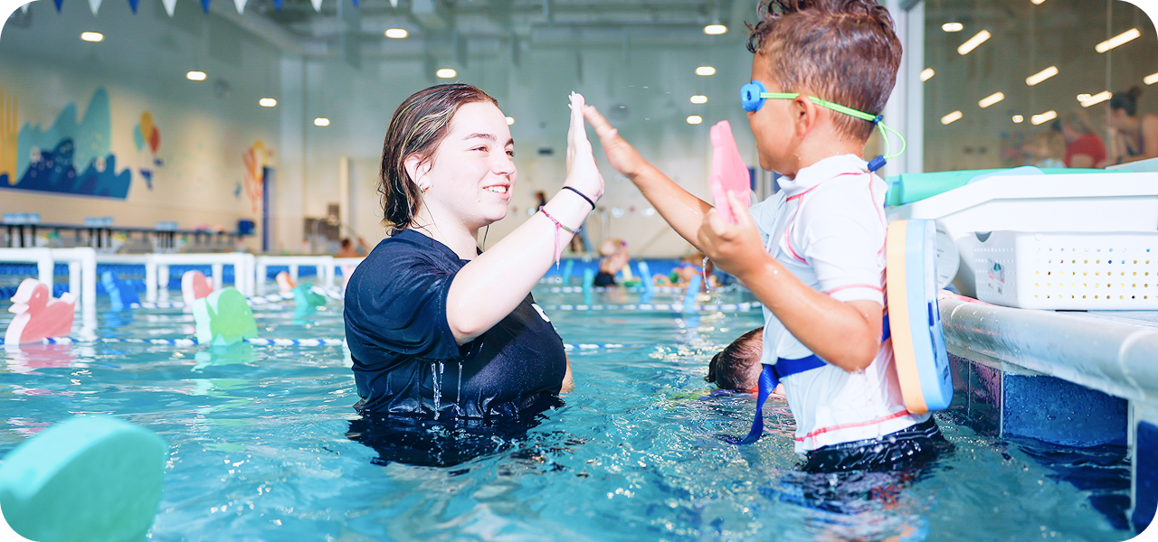 A young woman and a small boy in a swimming pool high-fiving each other. The woman is wearing a black t-shirt and the boy is wearing swim goggles and a rash guard. They are smiling and appear to be celebrating.