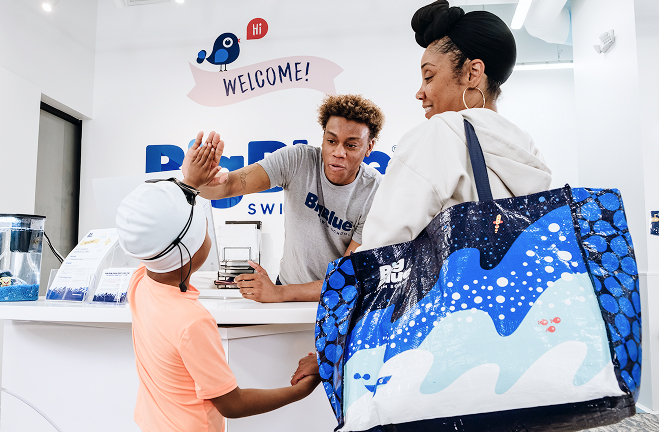 A young boy in a swim cap high-fives a swim instructor at a reception desk, while his mother stands beside them holding a large tote bag with a water-themed design.