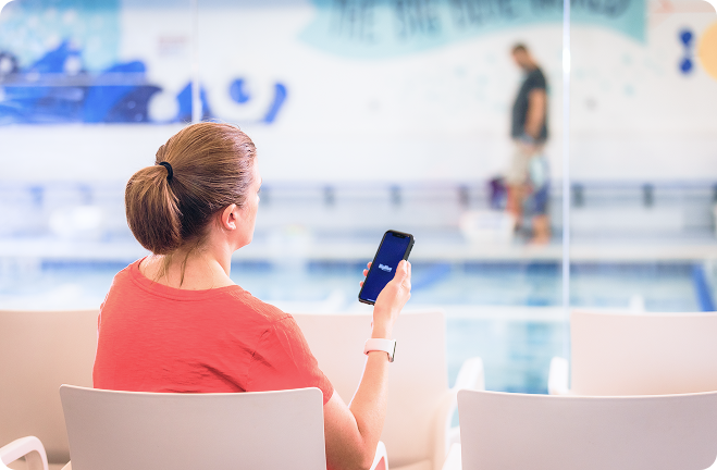 A woman in a red shirt holds a smartphone with a blue-themed app interface, seemingly looking at a swimming pool through a glass wall. She has her hair up in a ponytail and is wearing a white smartwatch.