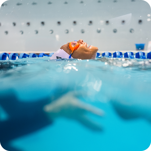 A swimmer with orange goggles and a white swim cap is seen floating on their back in a blue pool, face tilted up, arms and hands submerged below the water.