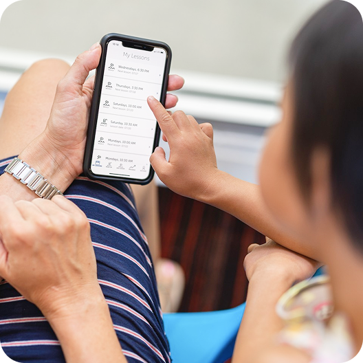 Person holding a smartphone displaying a "My Lessons" app interface, with a finger touching a lesson entry, and another person partially visible to the right.