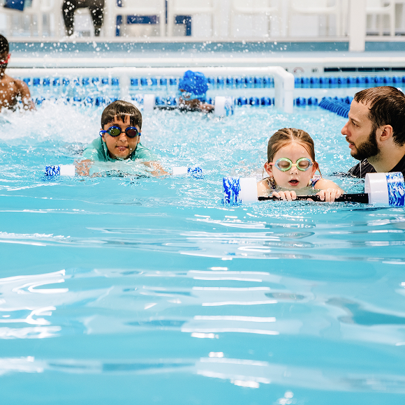 A group of children practices swimming with pool noodles in a bright indoor pool, water splashing around them.