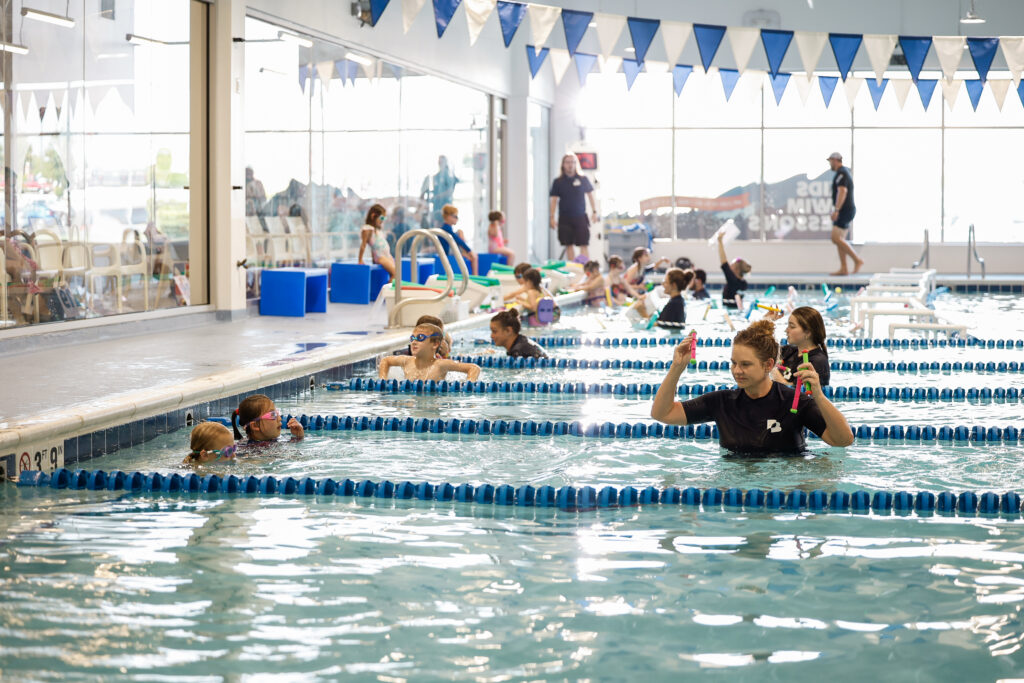 A swimming instructor demonstrates a technique while teaching a swim lesson to a group of children in an indoor pool, with blue lane dividers and blue triangle flags visible.