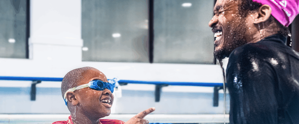 Young boy in blue goggles smiles and points towards an off-screen person, both appear wet from the swimming pool, as a laughing adult with a pink headscarf looks on.