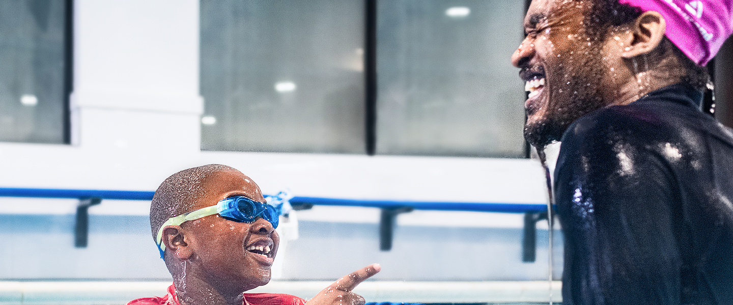Young boy in blue goggles smiles and points towards an off-screen person, both appear wet from the swimming pool, as a laughing adult with a pink headscarf looks on.
