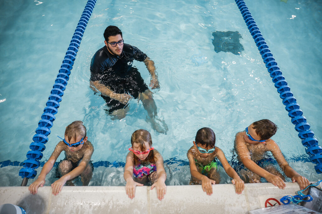 A swimming instructor guides four children in colorful swim gear at the edge of a pool, engaged in a lesson.