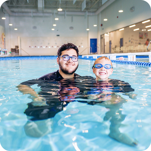 A young man with glasses and a small boy in goggles in a swimming pool, smiling and looking at the camera. They are both submerged up to their chests in the water.