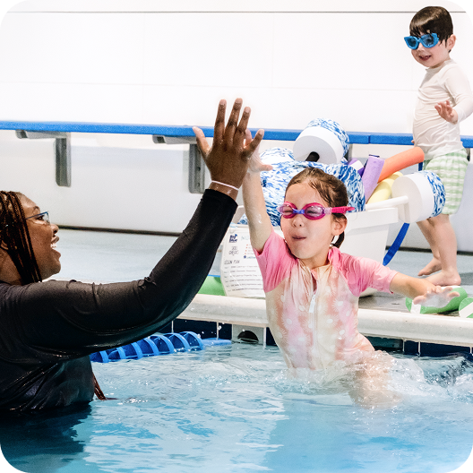 A young girl wearing pink swim goggles and a rash guard gives a high five to a swim instructor in a black long-sleeved swim shirt in a swimming pool, while another young child in swim goggles looks on.