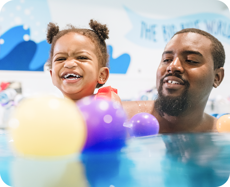 A young child in a colorful swimsuit enjoys playing with floating balls in a swimming pool, with playful wall art in the background.