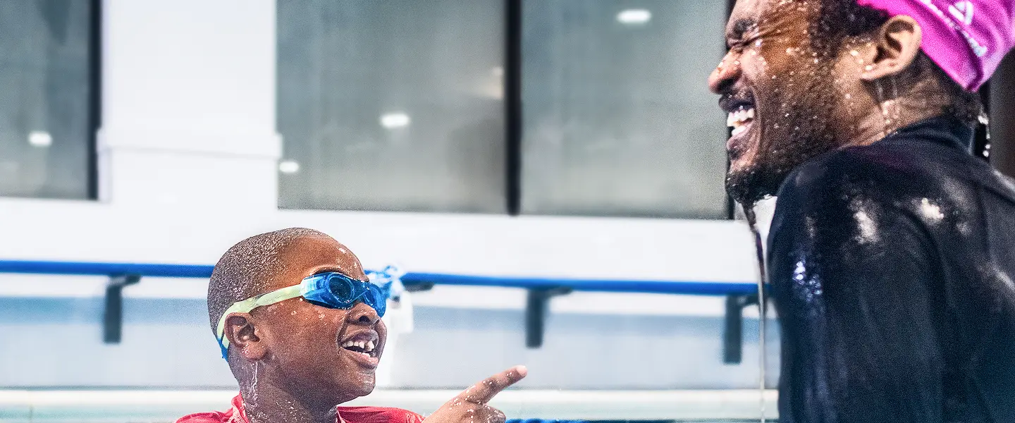 Young boy in blue goggles smiles and points towards an off-screen person, both appear wet from the swimming pool, as a laughing adult with a pink headscarf looks on.