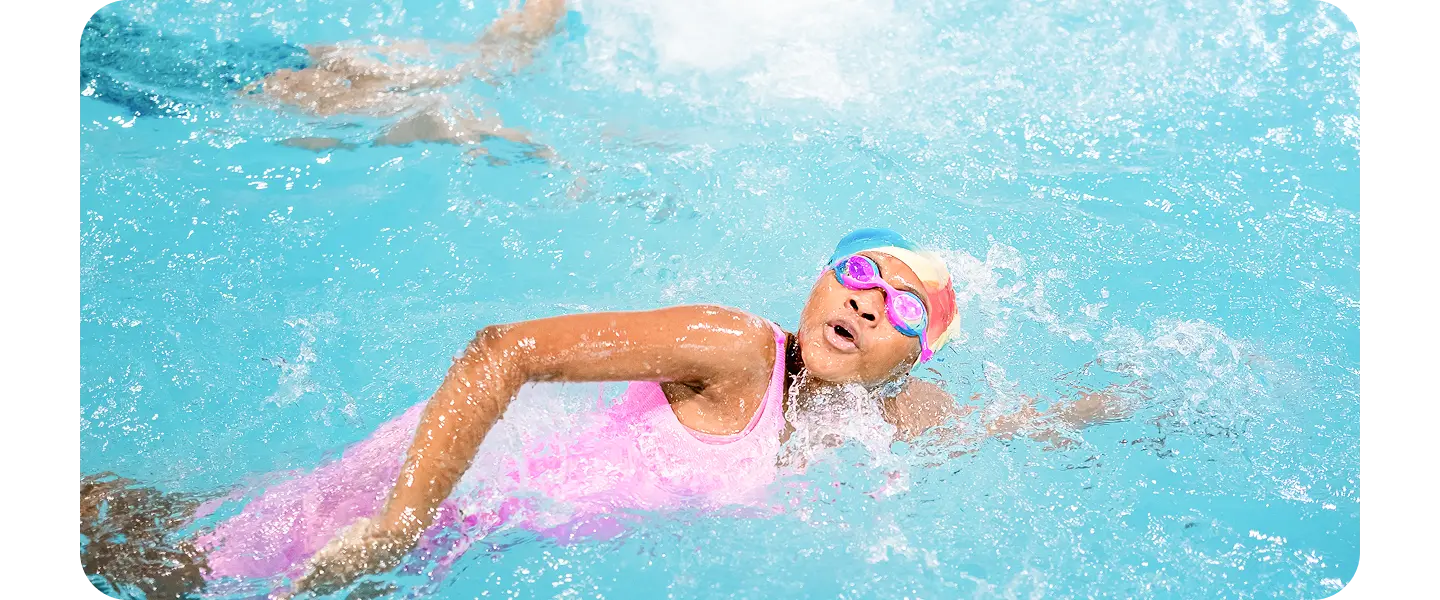 A young swimmer wearing a pink swimsuit, a multicolored swim cap and goggles, swims the front stroke in a pool.