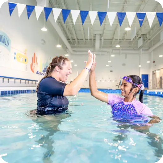 A swimming instructor and a young girl in the pool high-five, celebrating success, both smiling with joy, inside a brightly lit indoor pool facility with decorative pennants.