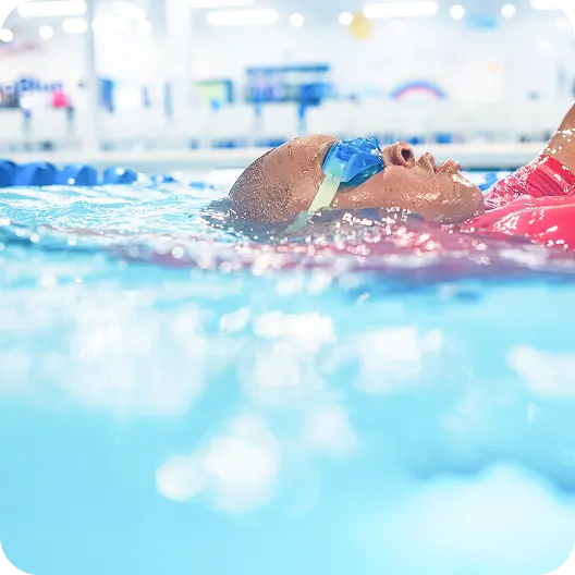 Person wearing blue swimming goggles floats on their back in a swimming pool, visible from the chest up, with bright blue water and pool interior in the blurred background.