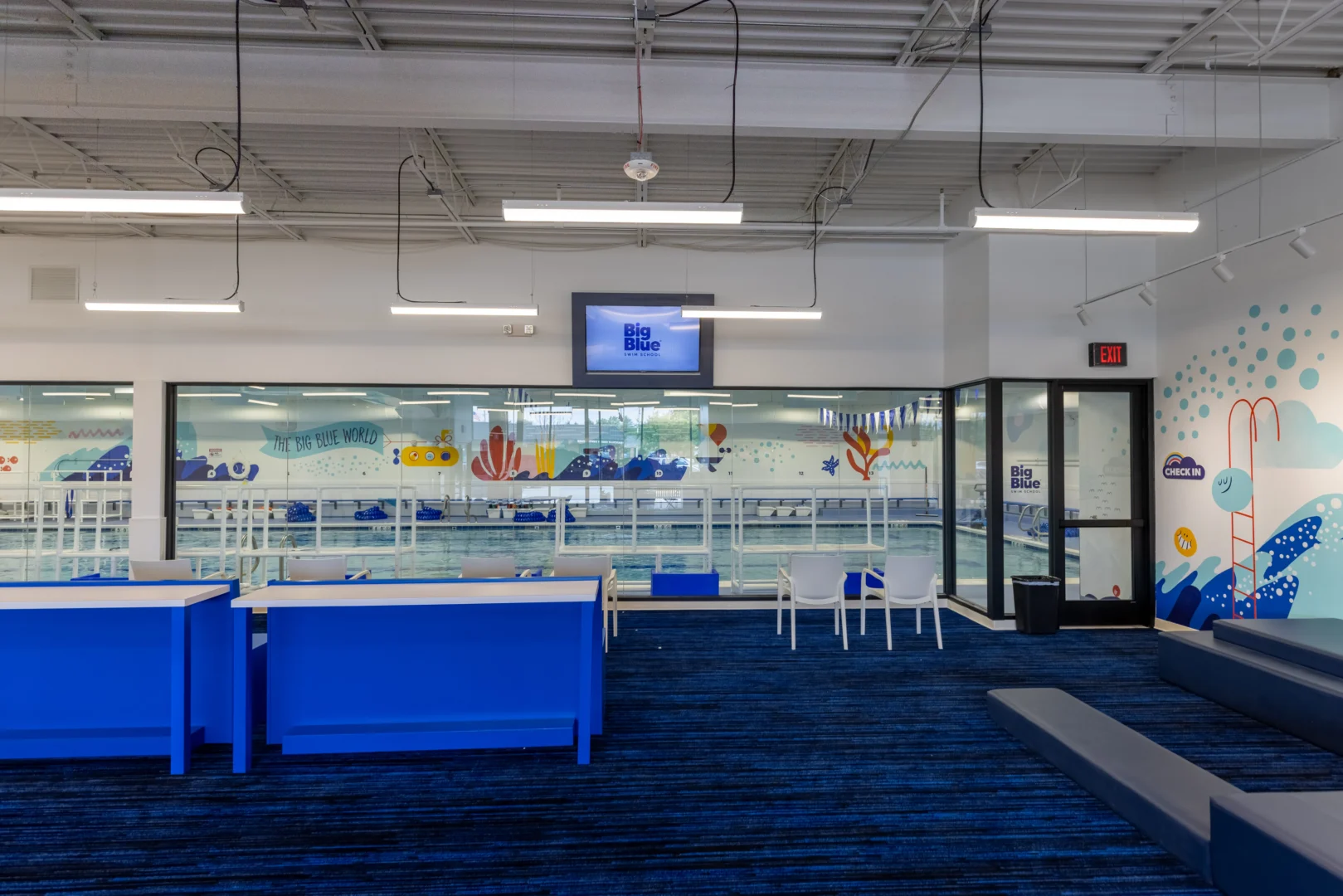 a waiting room with blue carpet and white chairs looking out onto a Big Blue Swim School pool area with colorful murals on the wall
