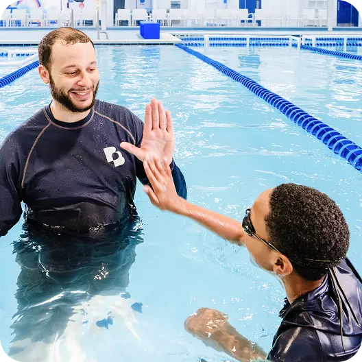 A man and a child in a swimming pool high-fiving, both wearing swim shirts and goggles, the man smiling.