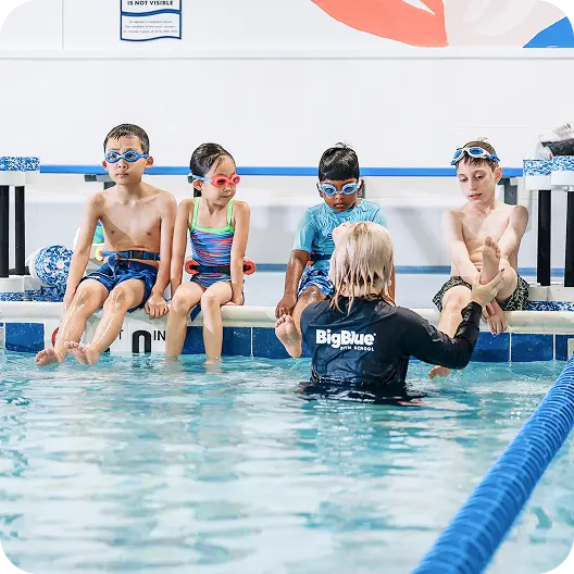 Four children in swim goggles and swimwear sit on the edge of a swimming pool as a swim instructor, wearing a Big Blue Bath School shirt, assists one child's foot in the water.