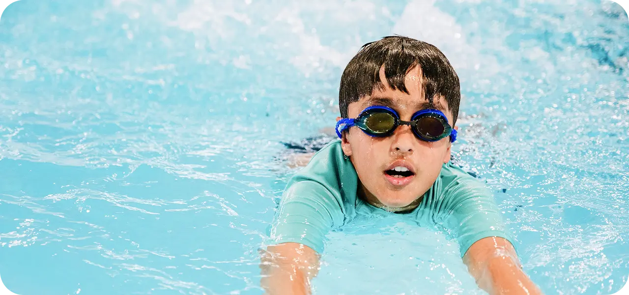 A young boy with dark hair and blue-tinted swim goggles swims freestyle in a pool, facing the viewer with an open mouth